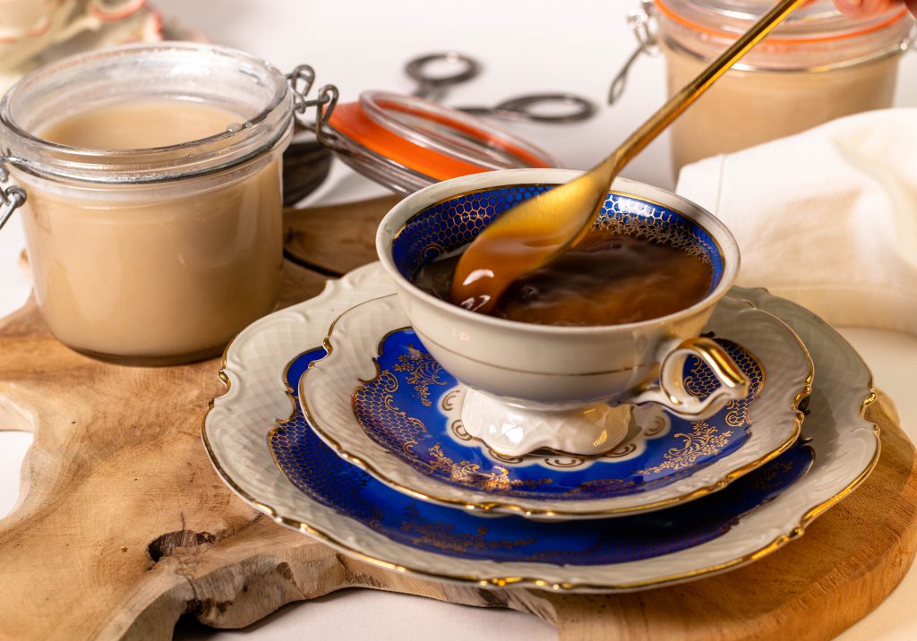 Zwei Gläser vegane Kondensmilch. Davor eine Kaffeetasse in der ein Löffel rührt.