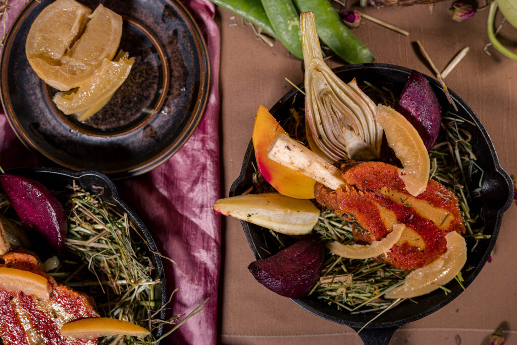 7. Die Scheiben der veganen Lammkarrees mit Salzzitronenschnitzen auf Heu und Ofengemüse portioniert in feuerfeste Formen geben.