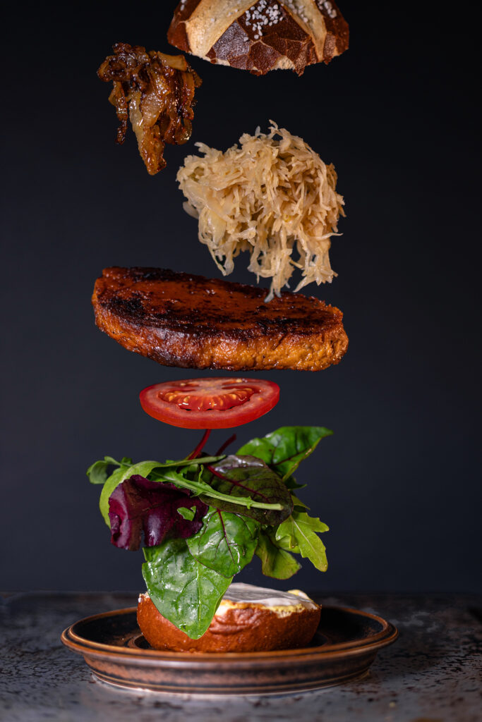 6. Die untere Burgerhälfte mit mittelscharfem Senf, süßem Senf und veganer Mayonnaise bestreichen. Dann mit Salat, Tomate, dem gebratenen Kasseler, Sauerkraut und Schmelzzwiebeln belegen.