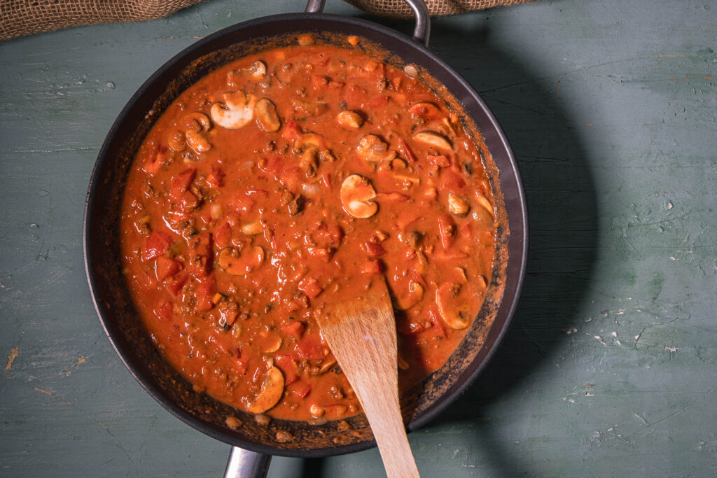 5. Mit dem Rotwein ablöschen. Dann die passierten Tomaten, das Tomatenmark, Wasser, vegane Sahne, Zucker, getrocknete und frische Tomaten einrühren. Etwa 15 Minuten köcheln.