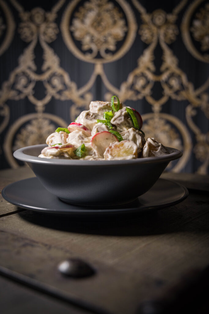 Gerösteter Kartoffelsalat in grauer Schale auf grauem Teller auf einer alten Truhe vor kitschiger Tapete.