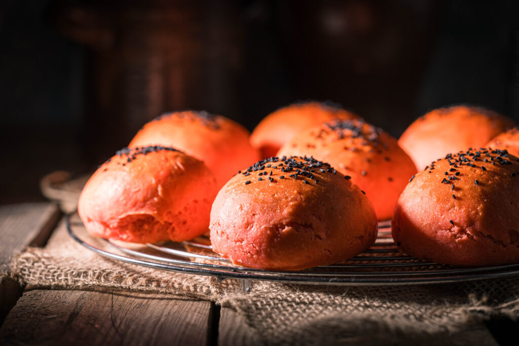 Rote Burgerbrötchen, bestreut mit Schwarzkümmel, auf einem Backrost auf einem Holztisch.