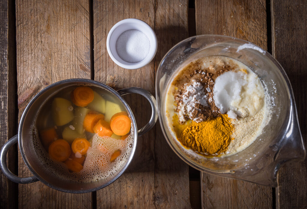 Möhrenscheiben und Kartoffelstücke in Wasser in einem kleinen Topf. Außerdem eine Schale Salz und ein Gefäss mit den restlichen Zutaten für die vegane Käsemasse.