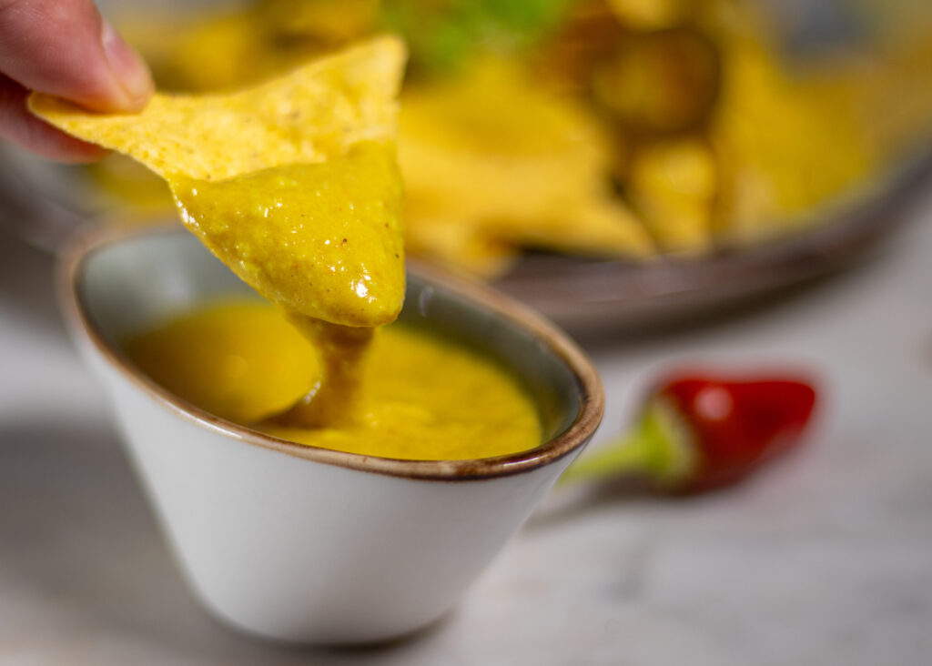 A Tortilla, being dipped into a vegan cheese dip. The cheese dip drops stringy from the tortilla.