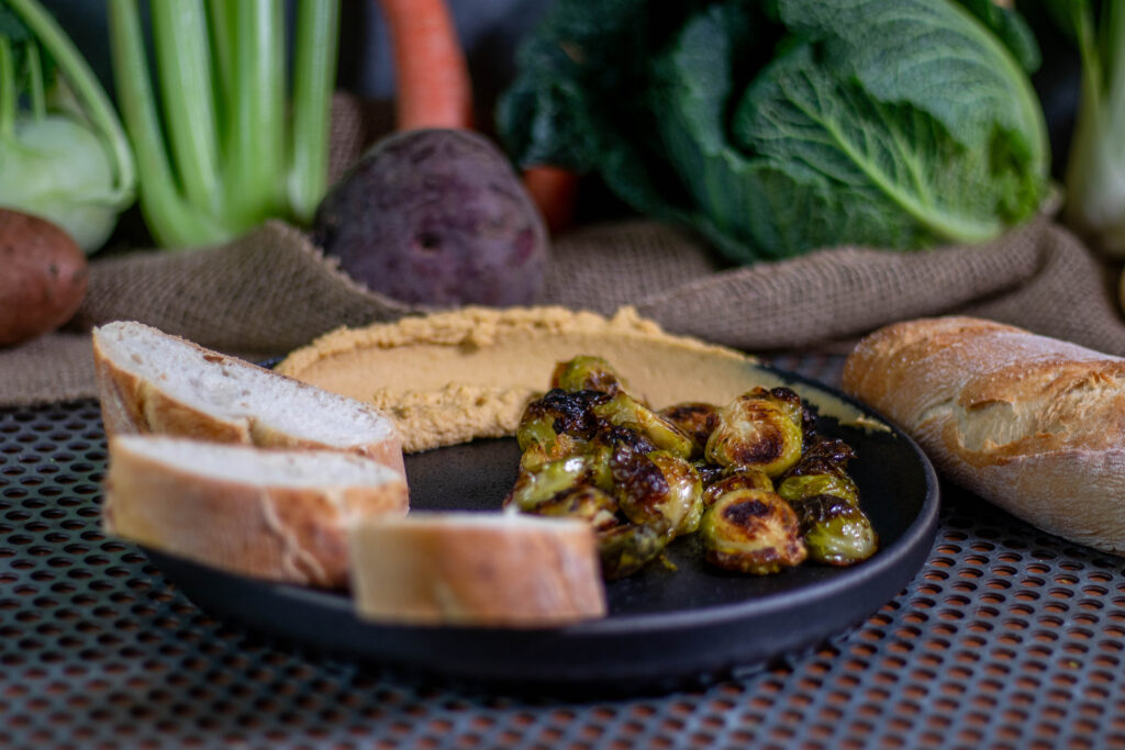 Oven roasted and glazed Brussel sprouts. With hummus and bread on a plate. Vegetables in the background.