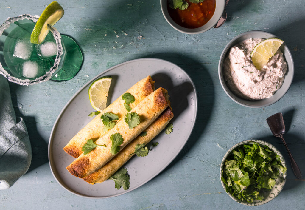 Drei vegane Chicken Taquitos von oben auf einem Teller. Außerdem auf dem Tisch: Schüsseln mit veganer Sour Cream, Koriander und Salsa, sowie ein Glas Magaritha.