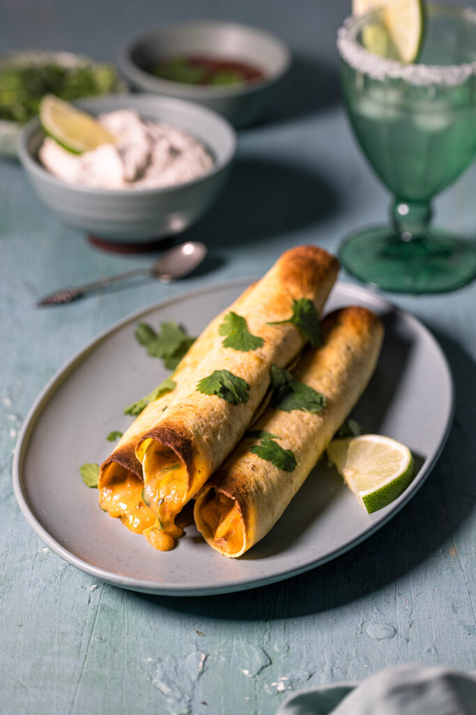 Drei vegane Chicken Taquitos auf einem Teller. Außerdem auf dem Tisch: Schüsseln mit veganer Sour Cream, Koriander und Salsa, sowie ein Glas Magaritha.