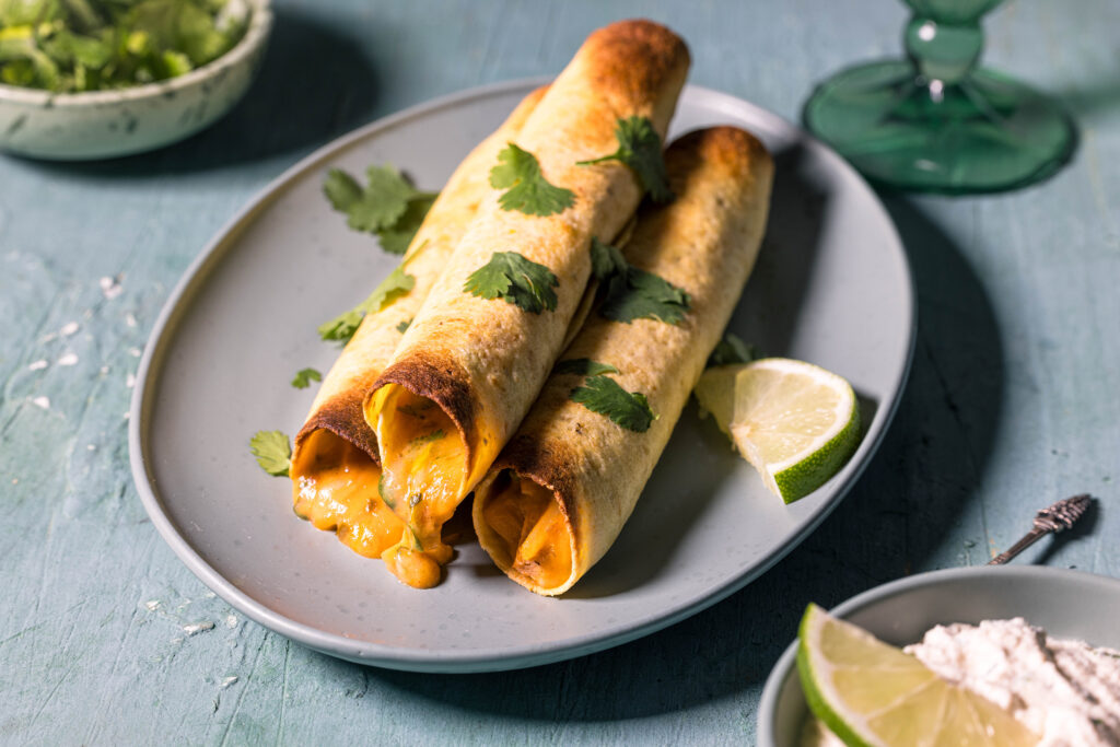 Drei egane, cremige Chicken Taquitos auf einem Teller. Die käsige Füllung läuft aus den Taquitos an einem Ende heraus.