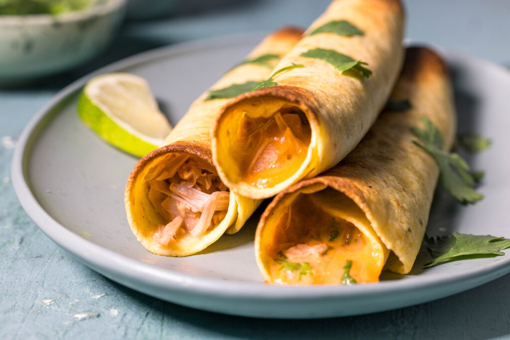 3 vegane Chicken Taquitos auf einem Teller. Daneben eine Limettenscheibe.