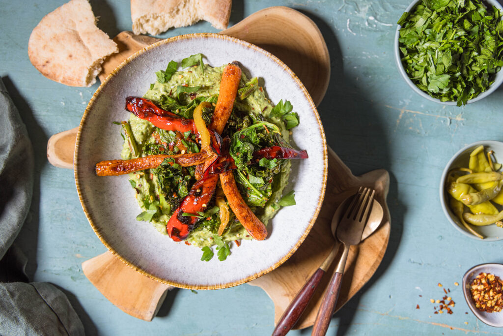 Gerösteter Grünkohl mit Ofengemüse auf Grünkohlhummus – in einer Bowl von oben fotografiert. Daneben Schälchen mit kleinen Beilagen.