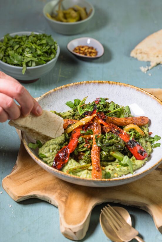 Gerösteter Grünkohl mit Ofengemüse auf Grünkohlhummus in einer Bowl. Eine Hand stippt Brot in den Hummus.