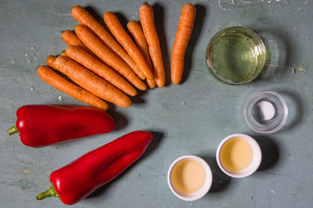 MInimöhren, Spitzpaprika und vier Schälchen mit Flüssigkeiten und Salz auf einem alten, grünlichen Tisch.