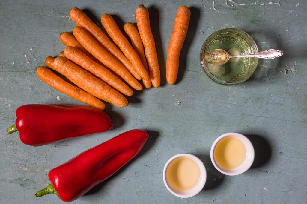 MInimöhren, Spitzpaprika und drei Schälchen mit Flüssigkeiten auf einem alten, grünlichen Tisch.