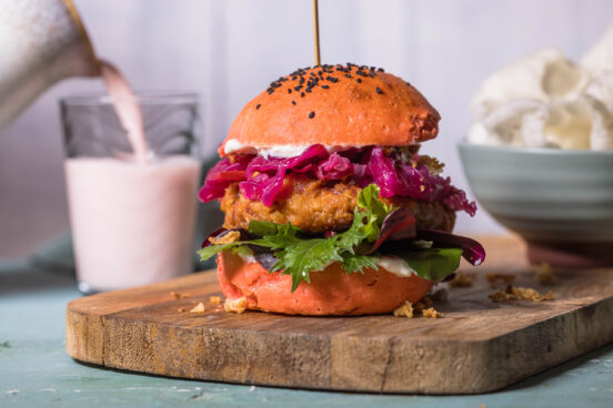 Veganer Kimchi-Burger auf Holzbrett. Im Hintergrund eine Schale Krupuk und ein Shake wird in ein Glas eingeschenkt.