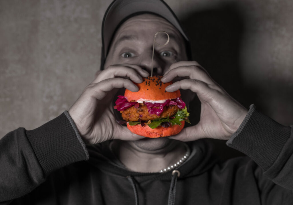 Sean Moxie beisst in einen Kimchi-Burger. Der Burger ist in Farbe, Sean Moxie schwarz-weiß.