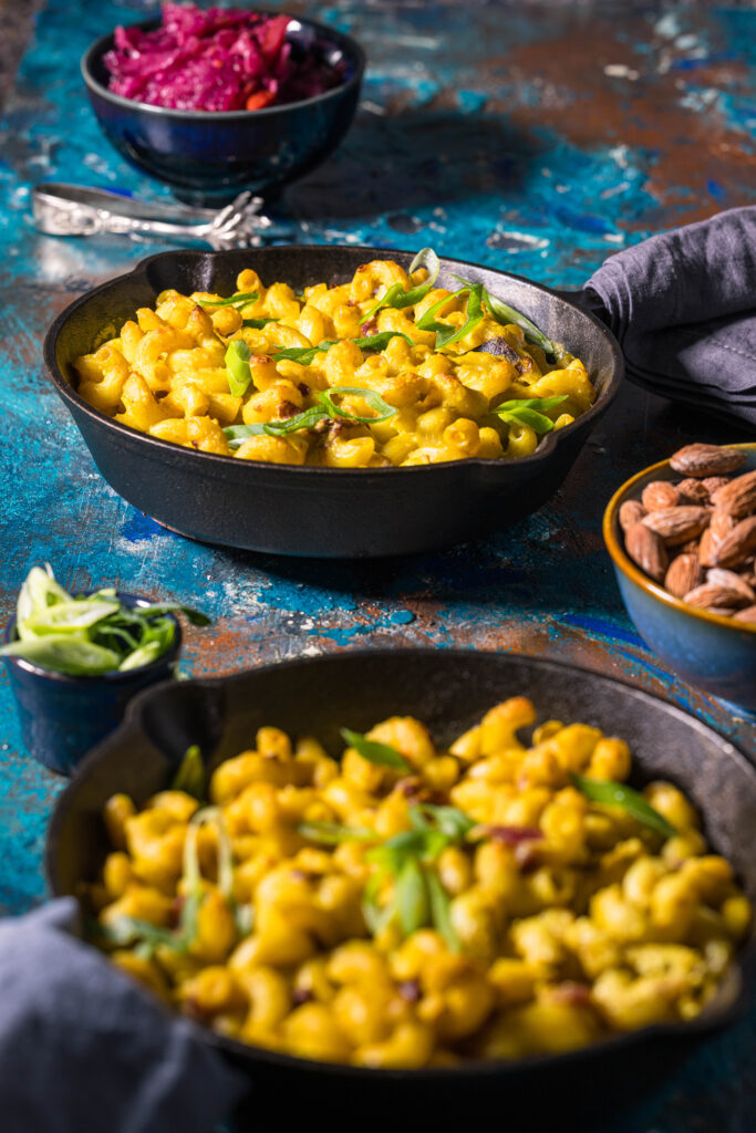 Veganes Kimchi Mac and Cheese in zweu kleinen gusseisernen Pfannen. Außerdem eine Schale mit Kimchi, eine mit Salzmandeln und eine mit Lauchzwiebeln auf einer bunten Oberfläche