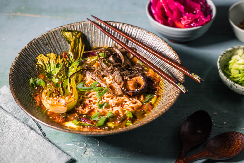 Kimchi Ramen in einer Bowl mit gebratenem Pak Choi und Nori. Daneben einige Schalen mit weiteren Toppings.