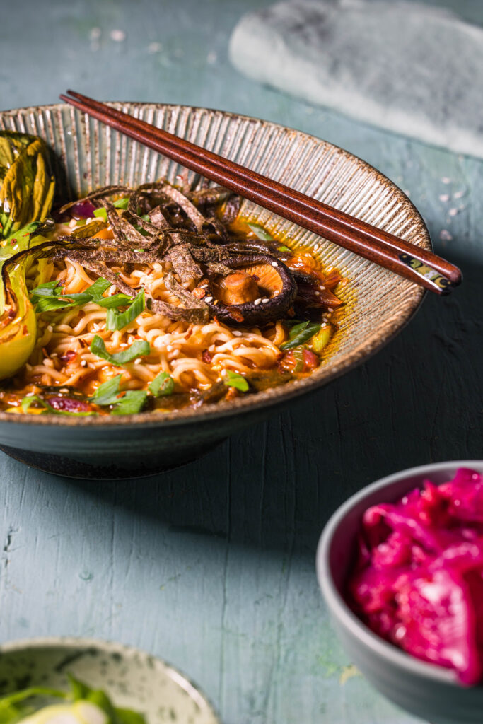 Koreanische Ramen, auch Ramyeon genannt, in einer Bowl mit Stäbchen. Davor eine kleine Schale mit Kimchi.