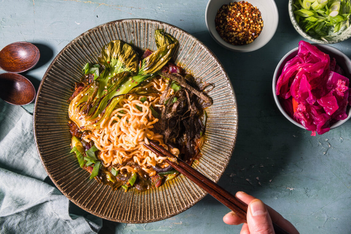 Vegane Kimchi Ramen mit gebratenem Pak Choi in einer großen Bowl, daneben Kimchi und weitere Toppings.