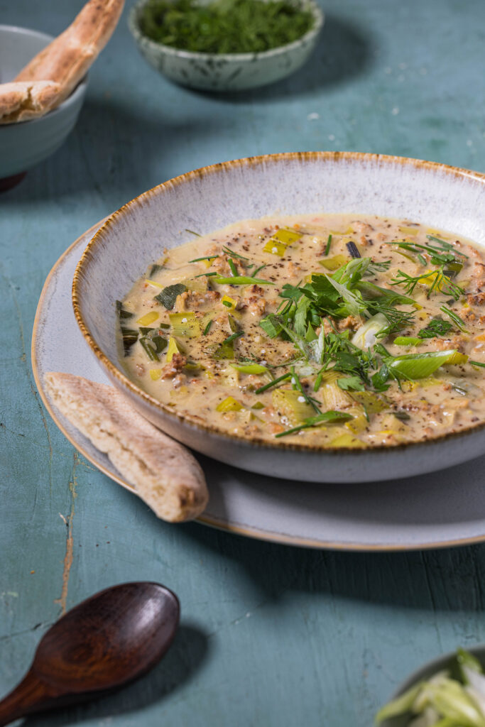 Vegane Lauch-Käse-Suppe mit Hack auf einem Teller mit Unterteller. Dazu Brot und eine Schale mit Kräutern.