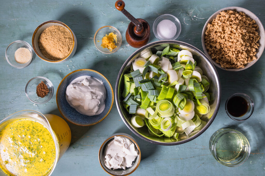 Alle Zutaten für die veganen Lauch-Hack-Käse-Suppe auf einem alten, grünlichen Tisch.