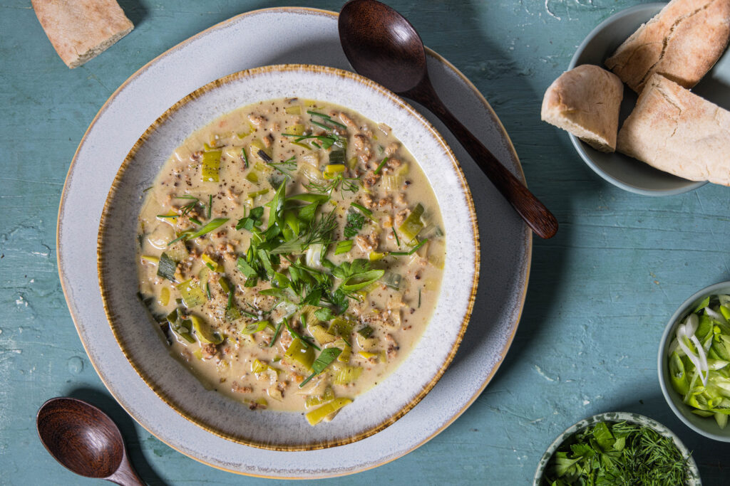 Vegane Lauch-Käse-Suppe mit Hack auf einem Teller mit Unterteller. Dazu Brot und eine Schale mit Kräutern und eine mit Lauchzwiebelringen.