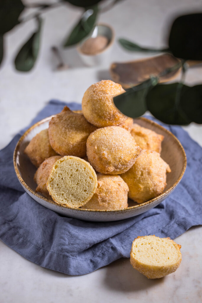 Vegane Quarkbällchen in einer Schüssel. Im Vordergrund ein verschwommener, grüner Zweig. Ein Quarkbällchen aufgeschnitten. Die zarte, luftige Krume des Teigs zeigt in die Kamera.