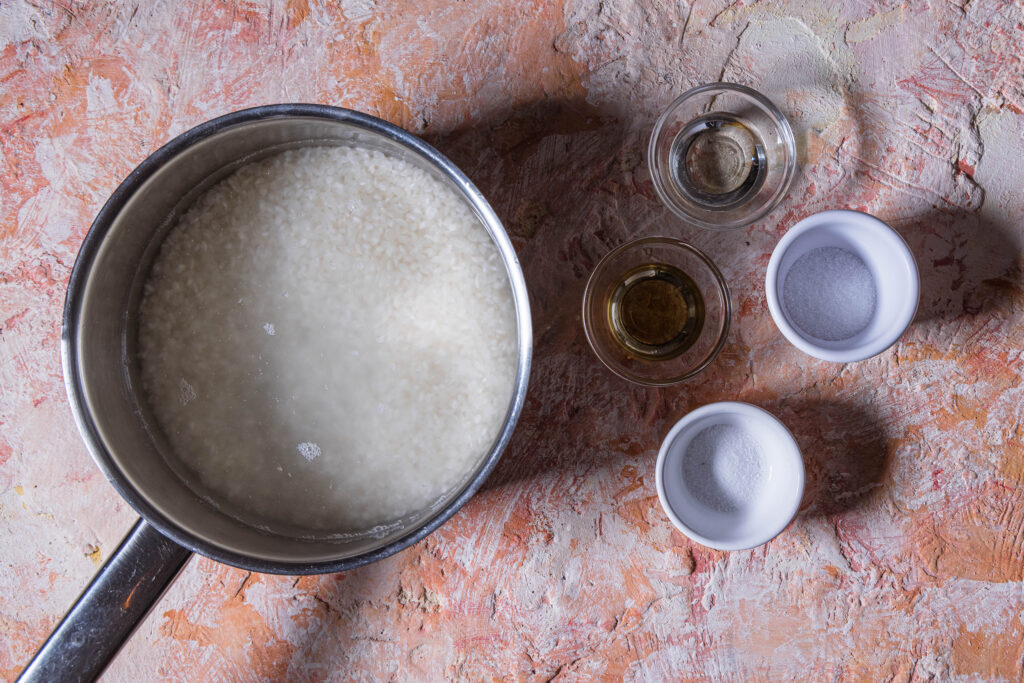 Sushireis mit Wasser in einem Topf. Weitere Zutaten in Schalen daneben.