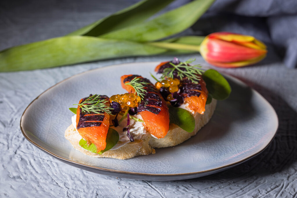 Selbstgemachter veganer Thunfisch aus Tomaten, auf Brot mit selbstgemachtem, veganen Kaviar. Hinter dem Teller liegt eine Tulpe.