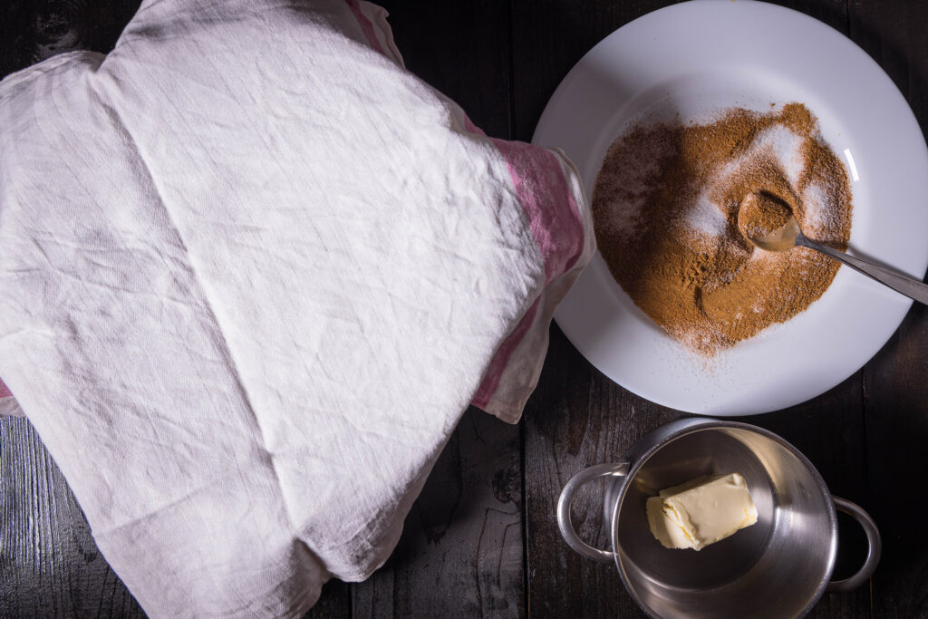 Eine Schüssel abgedeckt mit einem Geschirrtuch, daneben ein Teller mit Zucker und Zimt und ein kleiner Topf mit veganer Butter.