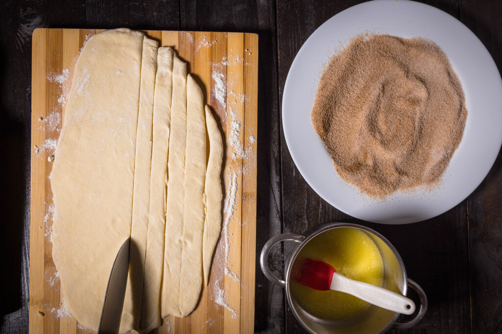 Ausgerollter Hefeteig auf einem Holzbrett wird in schmale Streifen geschnitten. Daneben Zucker und Zimt auf einem Teller und geschmolzene, vegane Butter in einem Topf.