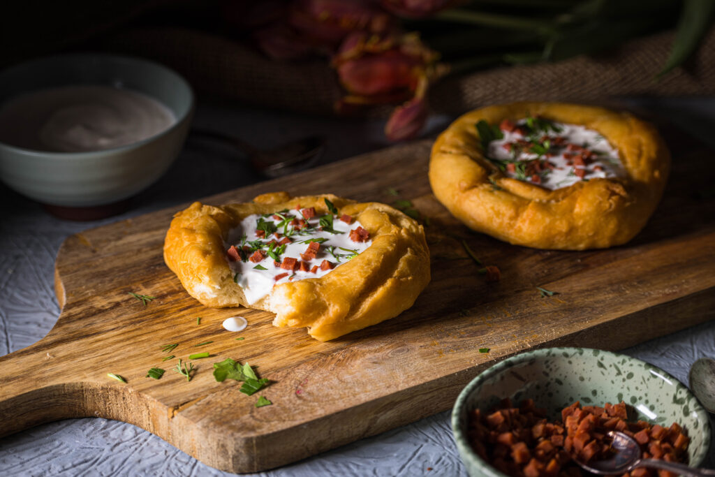 2 vegane Lángos auf einem Holzbrett. Davor und dahinter kleine Schalen mit Toppings.