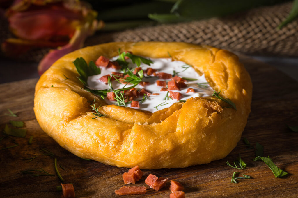 Ein frischer Lángos auf einem Holzbrett. Mit veganem Sauerrahm, Speck und Kräutern als Toppings.