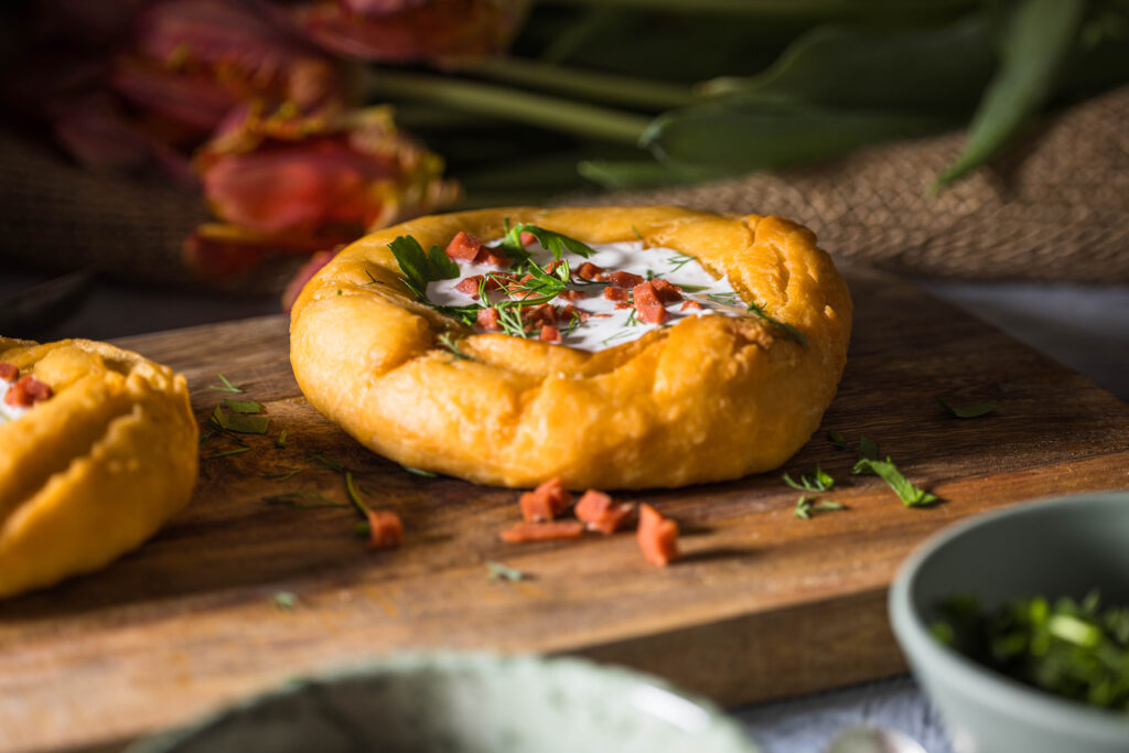 2 vegane Lángos auf einem Holzbrett. Davor und dahinter kleine Schalen und ein Strauss Tulpen.