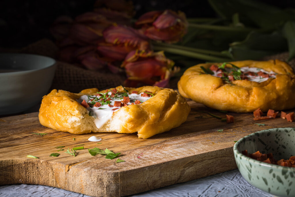 2 vegane Lángos auf einem Holzbrett. Davor und dahinter kleine Schalen und ein Strauss Tulpen.