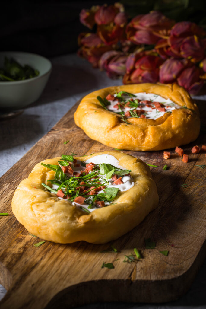 2 vegane Lángos auf einem Holzbrett. Dahinter kleine Schalen und ein Strauss Tulpen.