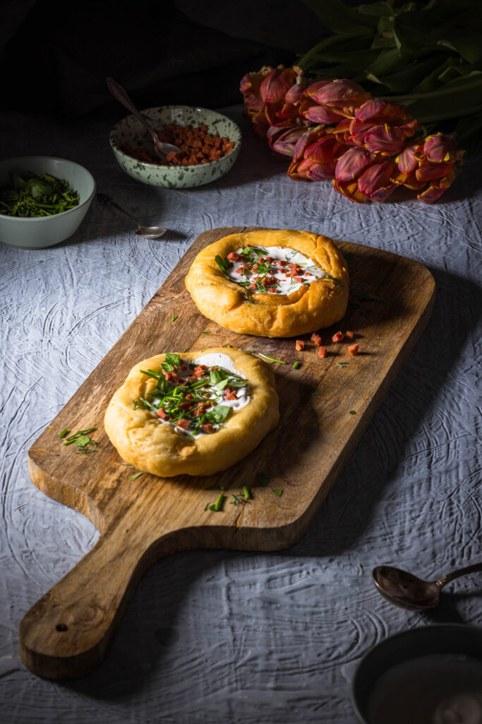 2 vegane Lángos auf einem Holzbrett. Dahinter kleine Schalen und ein Strauss Tulpen.