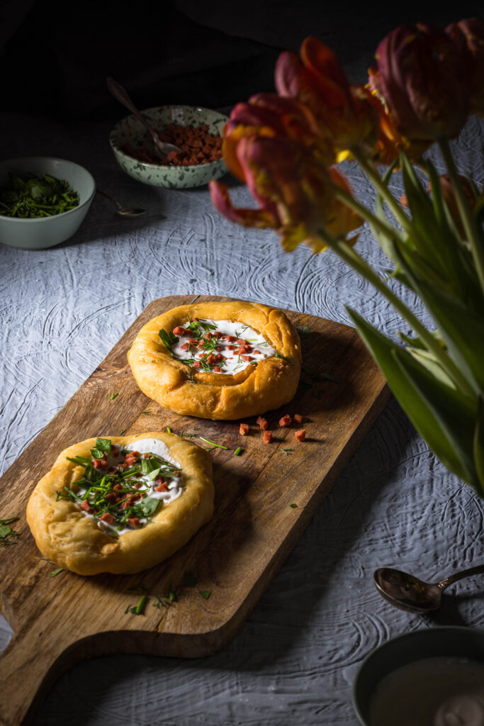 2 vegane Lángos auf einem Holzbrett. Davor ein Strauss Tulpen. Dahinter kleine Schalen.