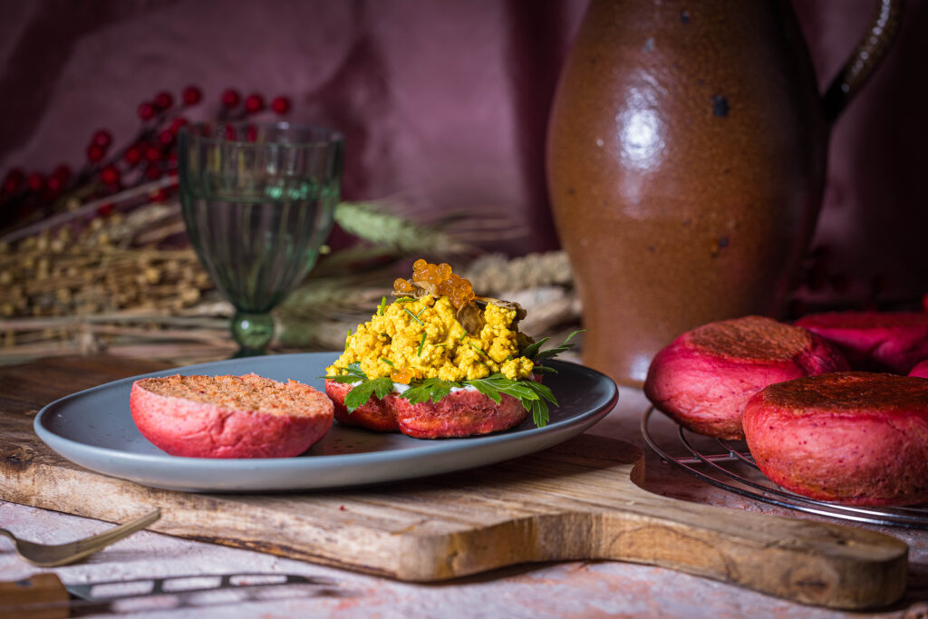 Rosa Rote Beete Pfannenbrötchen, aufgeschnitten, eine Hälfte ist mit Scrambeld Tofu, veganem Kaviar und eingelegten Pilzen belegt. Daneben mehr Pfannenbrötchen.