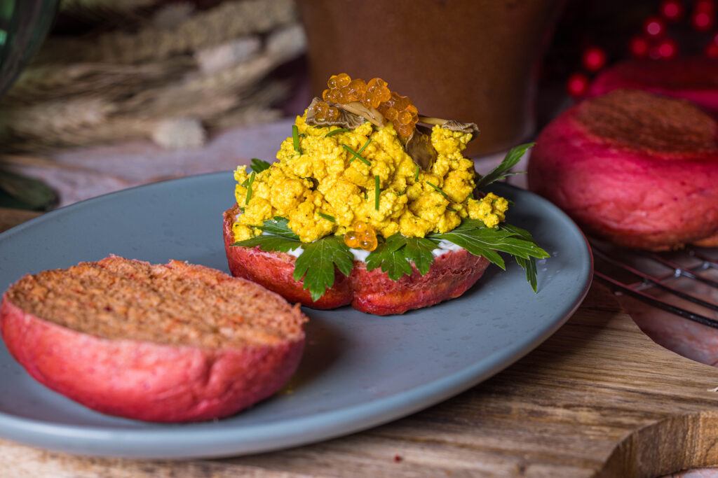 Rosa Rote Beete Pfannenbrötchen, aufgeschnitten, eine Hälfte ist mit Scrambeld Tofu, veganem Kaviar und eingelegten Pilzen belegt. Im Hintergrund mehr Pfannenbrötchen.