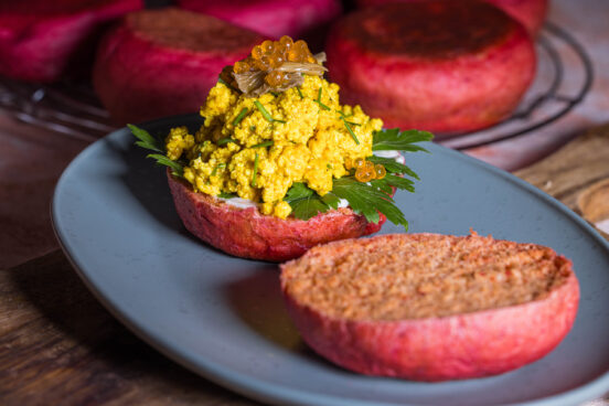 Rosa Rote Beete Pfannenbrötchen, aufgeschnitten, eine Hälfte ist mit Scrambeld Tofu, veganem Kaviar und eingelegten Pilzen belegt. Im Hintergrund mehr Pfannenbrötchen.