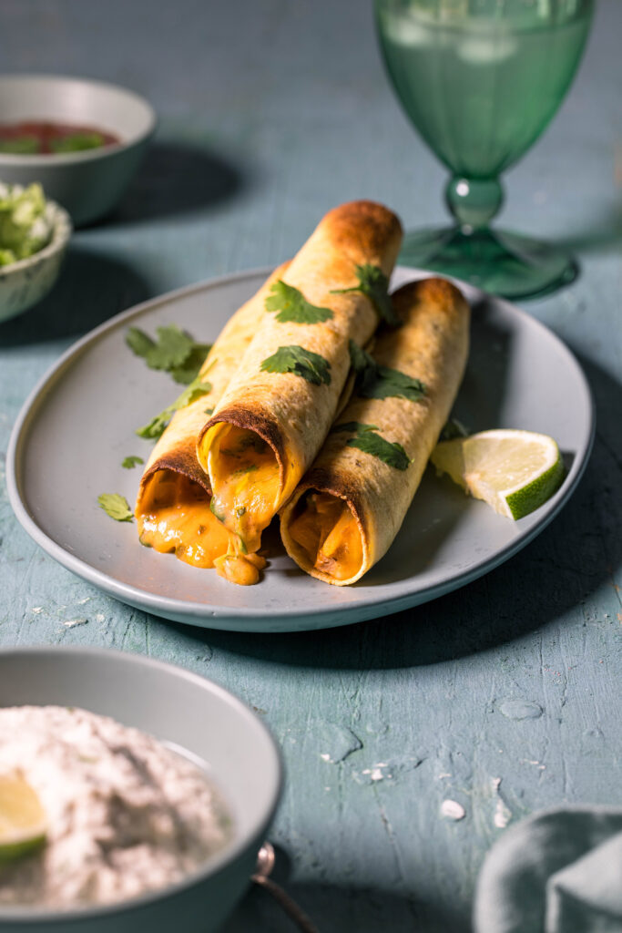 Drei Taquitos auf einem Teller. Davor eine Schale vegane Sour Cream.