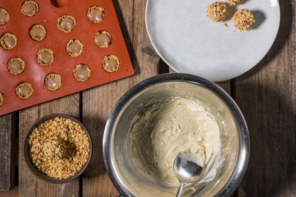 A silicone mold with half filled Viottos, a plate with finished Giottos, a bowl with chopped hazelnuts in which a Giotto is being turned, and a bowl with melted white chocolate.